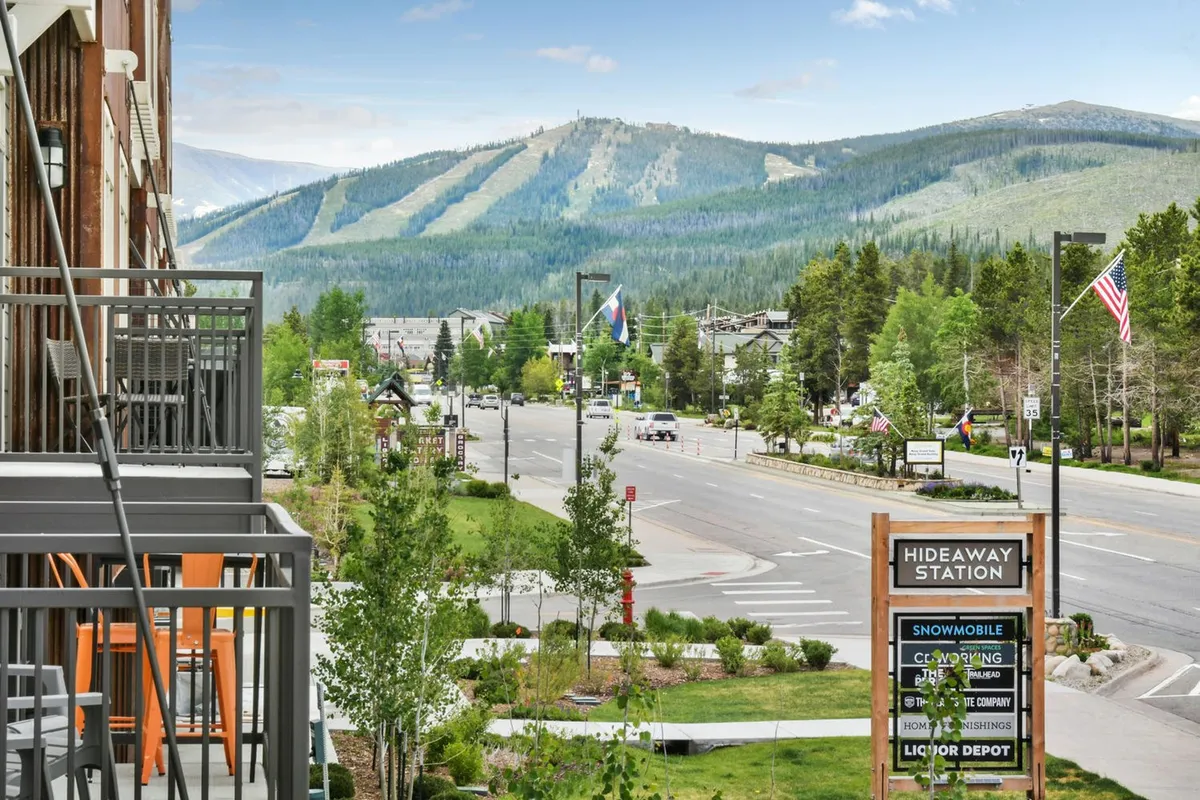 This exterior image frames a balcony with seating, magnificent mountain views, and the nearby Hideaway Station sign, linking the property to the vibrant local scene and hinting at easy access to year-round attractions.