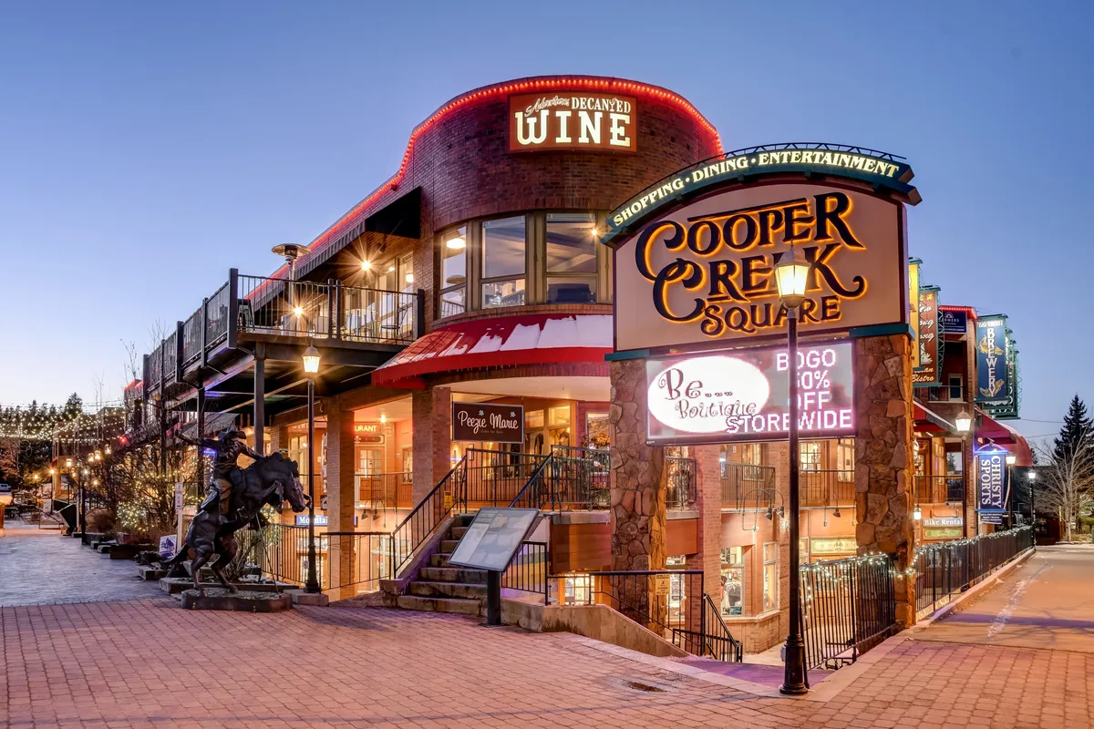 A vibrant exterior view of Cooper Creek Square, featuring a welcoming sign, decorative string lighting, and visible entrances to dining and shopping venues. The scene is alive with the potential for social interaction, entertainment, and culinary exploration.
