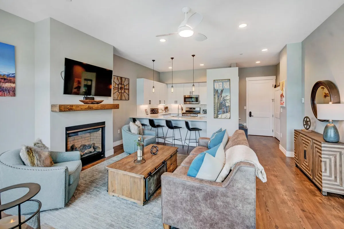 The image showcases a stylish and cozy open-concept living area with a glowing fireplace, plush sofa, modern kitchen island, and tasteful artwork on the walls. The setting invites guests to relax by the fire, gather for lively conversations, or enjoy casual meals together, all within a beautifully designed space.