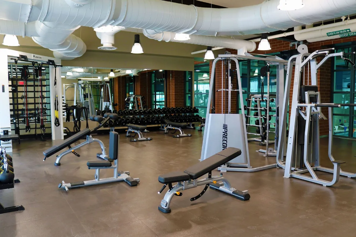 A lively fitness center scene featuring weight lifting equipment, exercise benches, mirrored walls, and dumbbell racks. The space is filled with natural light and feels energetic, motivating, and community-oriented.