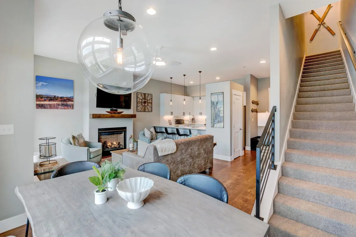 A welcoming interior featuring a dining table adorned with plants, a cozy seating area next to a fireplace, and a staircase leading to another part of the home. The space is styled for comfort and socializing, with ample natural light and thoughtful touches.