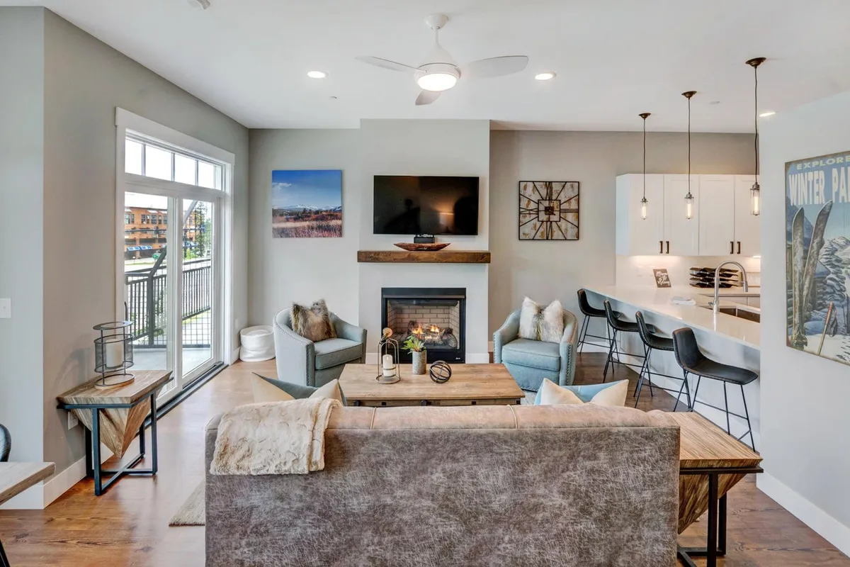 A modern living area centered around a programmable gas fireplace, with a cozy sofa facing a sleek HD smart TV. Large windows fill the space with sunlight, and bar stools sit adjacent to the main seating area, hinting at the easy transition between relaxation and dining.