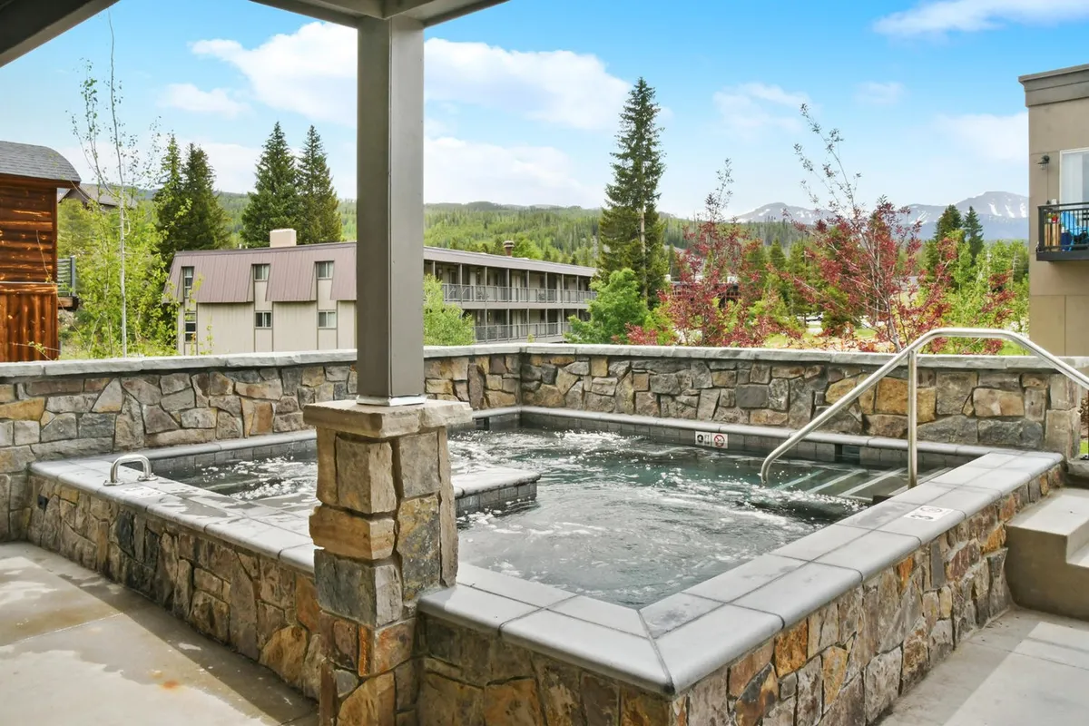 A large, inviting hot tub sits outdoors surrounded by nature, with mountains forming a dramatic backdrop. Guests are shown unwinding in the hot tub, emphasizing its impressive size and communal, restorative nature after adventure-packed days.