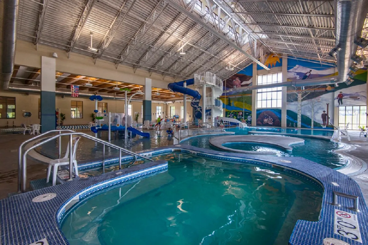 Inside a recreation center, children play joyfully in a vibrant water park setting complete with a colorful waterslide, wave pool, and murals. The atmosphere is energetic and filled with family activity, highlighting swimming and water play.