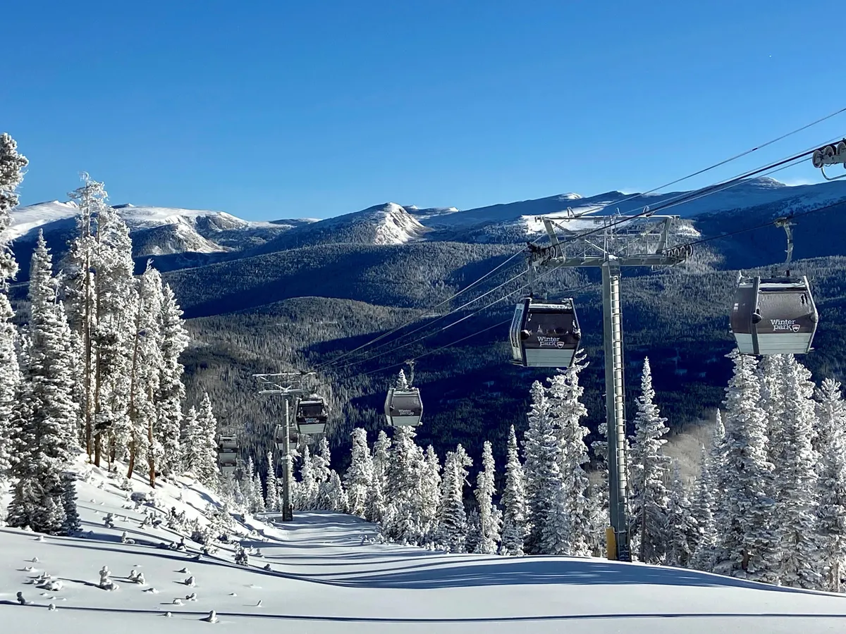 A panoramic view of ski lift gondolas gliding above snow-covered trees and towering mountain ranges, with hints of morning light or dusk creating a tranquil, majestic scene. The image exudes the anticipation and beauty of access to Winter Park Resort's lifts.